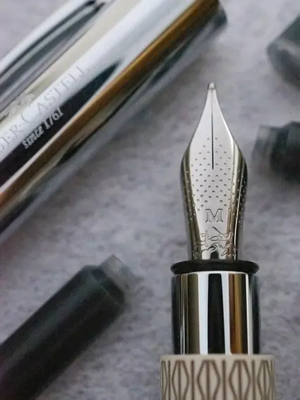 Eine Nahaufnahme einer Füllerfeder der Stärke M. Der silberne Füller von Faber-Castell liegt auf einem grauen Untergrund. Drumherum sieht man im Anschnitt mehrere schwarze Tintenpatronen, sowie den silbernen Deckel des Füllers. Foto: Johanna Rundel