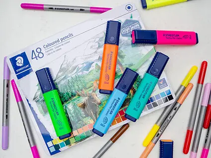 Verschiedene Fasermaler, bunte Textmarker und ein Blechetui mit Buntstiften der Marke Staedtler liegen verstreut auf einem betongrauen Untergrund.