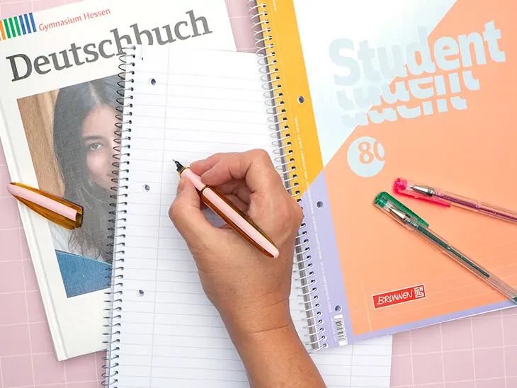 Jemand schreibt in einen linierten Collegeblock von Brunnen. Daneben liegt ein Deutschbuch für die weiterführende Schule. Foto: Johanna Rundel