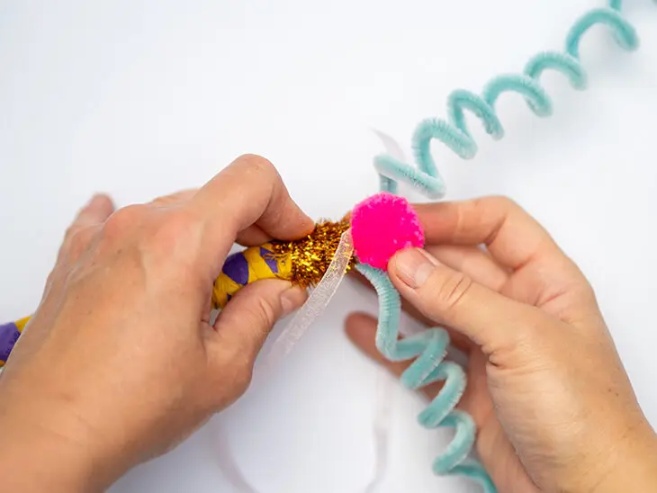 Jemand klebt einen neonpinken Pompon von KNORR prandell auf einen mit Materialien von HEYDA verzierten Zauberstab aus einem Ast. Foto: Johanna Rundel