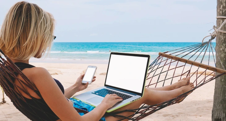 Frau arbeitet entspannt mit Laptop und Smartphone in Hängematte am Strand.