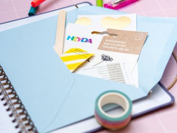 Ein hellblauer Briefumschlag ist innen auf die Rückseite eines Schülerkalenders geklebt. Daraus schauen verschiedene Stickerbögen und Materialien von HEYDA heraus. Foto: Johanna Rundel