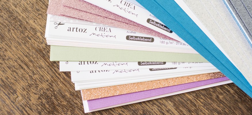 Eine Auswahl an verschiedenen Bastelpapieren von Artoz liegt aufgefächert auf einem Holztisch.
