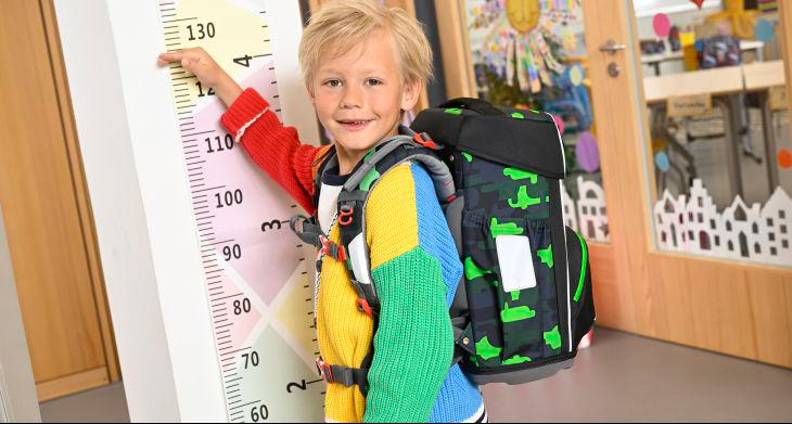 Ein Junge mit einem Schulranzen von Step by Step auf dem Rücken steht vor einer Messlatte und hält seine Hand daran