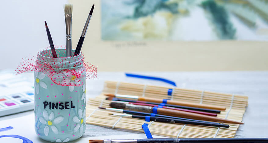 Vor einem unscharfen Hintergrund steht ein upcyceltes Marmeladenglas mit Malpinseln. Das Glas ist hellblau. Darauf sind weiße Blumen und rosa Punkte gemalt. Auf dem Glas steht in schwarzer Schrift "Pinsel". Über der Glasöffnung ist ein rotes Netz gespannt, in dem die Pinsel stecken. Um das Glas sind verschiedene Malutensilien verteilt. Unter anderem eine Bambusmatte, in der Pinsel aufgerollt werden können.