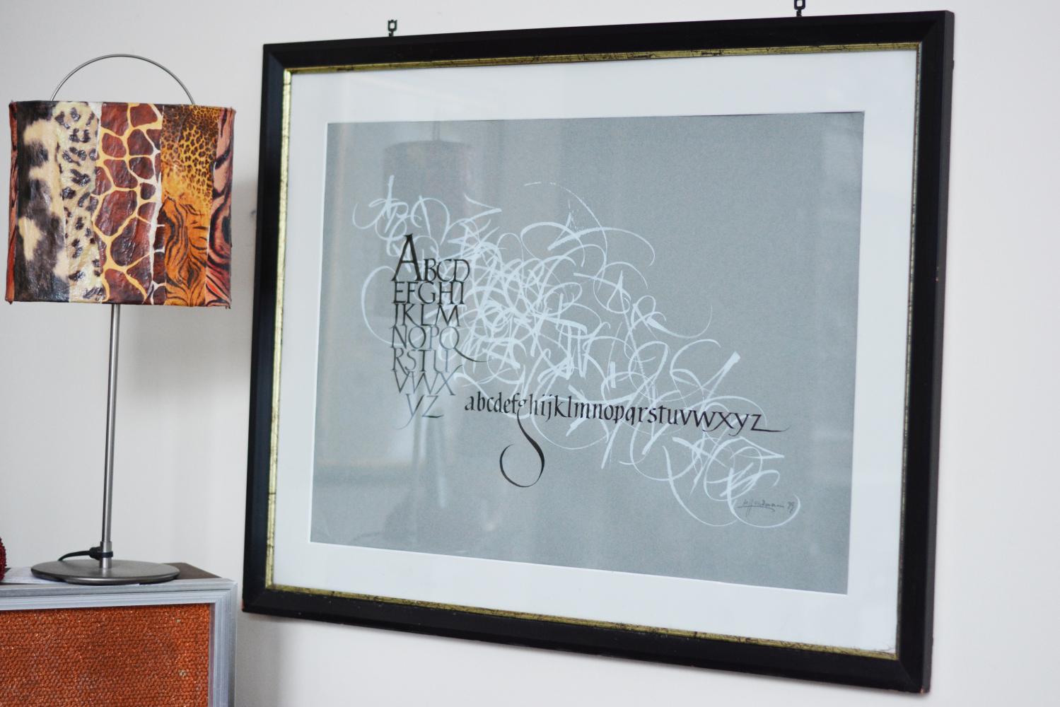 Ein Bild mit einem kalligrafierten Alphabet hängt in einem schwarzen Holzbilderrahmen an der Wand. Neben dem Bild ist eine Tischleuchte auf einem Sideboard zu sehen.