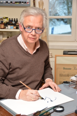 Martin Heickmann sitzt in seinem Museum am Schreibtisch. Er schaut in die Kamera, hält eine Kalligrafie-Feder in der Hand und kalligrafiert etwas auf einem Blatt Papier.