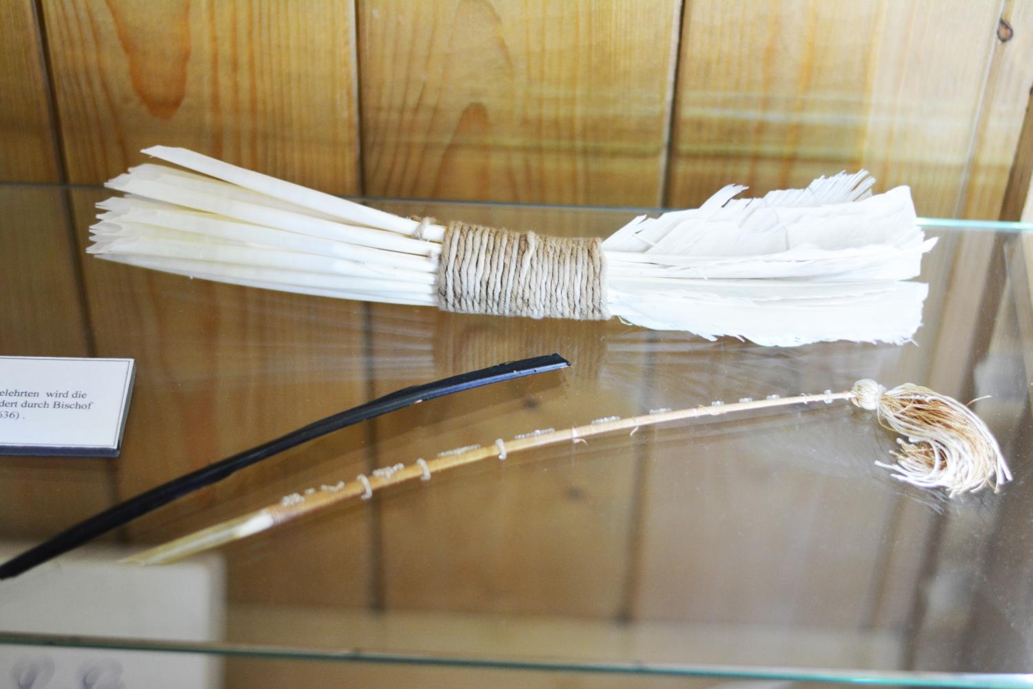 Auf einer Glasplatte einer Vitrine liegen weiße Schreibfedern zusammengebunden, daneben liegt eine einzelne schwarze und eine einzelne braune Feder, welche ein Troddel am Ende der Feder hat.