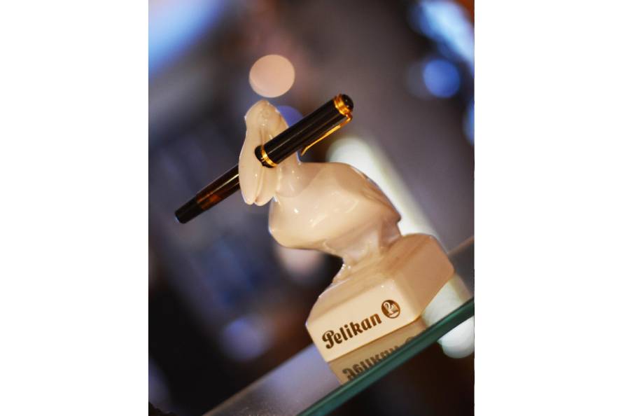Porzellanfigur eines Pelikans, der einen Pelikan Füller hält. Auf dem Sockel ist das Pelikan Logo aufgedruckt.