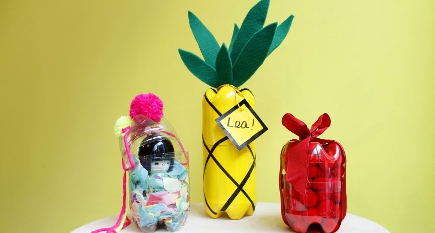 Vor einem gelben Hintergrund stehen 3 gebastelte Geschenkboxen aus Plastikflaschen, eine in Form einer Ananas mit einem Anhänger mit dem Namen "Lea", eine durchsichtige PET-Flasche, in die eine Spielfigur und bunte Papierschnipsel gefüllt sind. Die Flasche ist mit einem pinken und gelben Bommel verzeiert. Die dritte PET-Flasche ist ist rot und mit einer roten Schleife verziert.