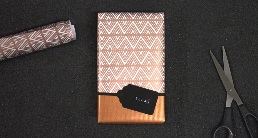 Auf einem schwarzen Untergrund liegt ein quadratisches Geschenk, welches in bronzefarbenes Geschenkpapier eingepackt ist. Auf dem Papier klebt ein Geschenkanhänger mit dem Namen Ella drauf. Daneben liegt eine Schere und ein Rest des Geschenkpapiers.