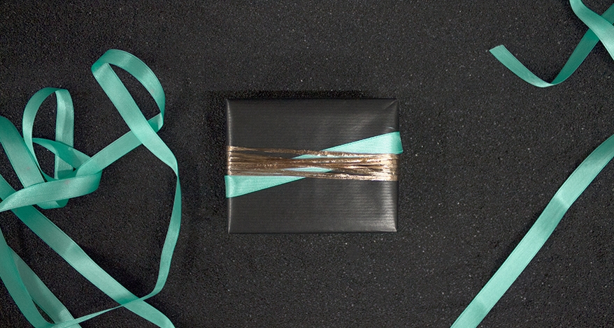 Auf einem schwarzen Untergrund liegt ein quadratisches Geschenk, welches in schwarzes Geschenkpapier eingepackt ist. Das Geschenk ist mit goldenem und türkisem Schleifen- und Satinband umwickelt. Daneben liegt ein Rest des türkisen Satinbands..