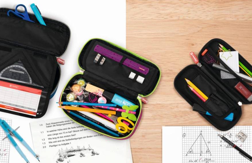 Auf einem Tisch liegen Blätter und 3 geöffnete Schlampermäppchen, gefüllt mit Geometrie-Dreick, Stiften und Schere.