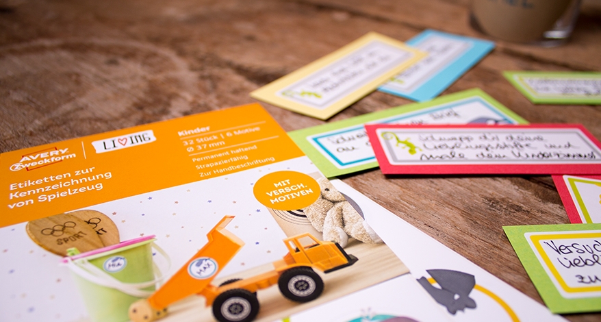 Nahaufnahme der Verpackung der Avery Zweckform Living Etiketten zur Kennzeichnung von Spielzeug. Daneben liegen bunte Aufgaben-Kärtchen, welche mit den Stickern beklebt sind.