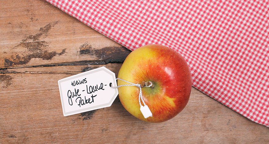 Ein Apfel liegt auf einem Holztisch neben einem rot-weiß karierten Tischtuch. Ane dem Apfel hängt ein Tafel-Etikett mit der Aufschrift "Kleines Gute-Laune-Paket".