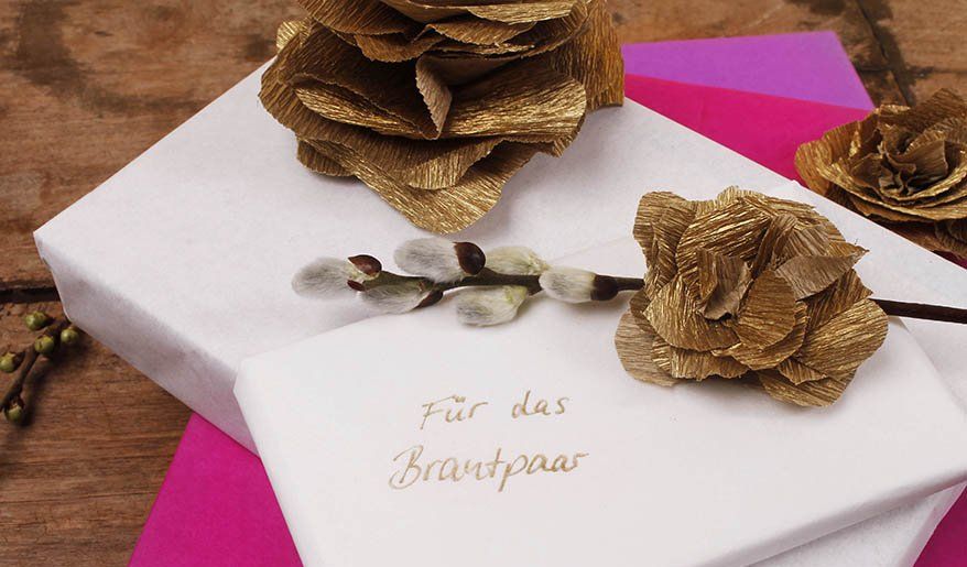 Goldene Blumen aus Krepppapier liegen als Deko auf 2 Geschenken für ein Brautpaar.