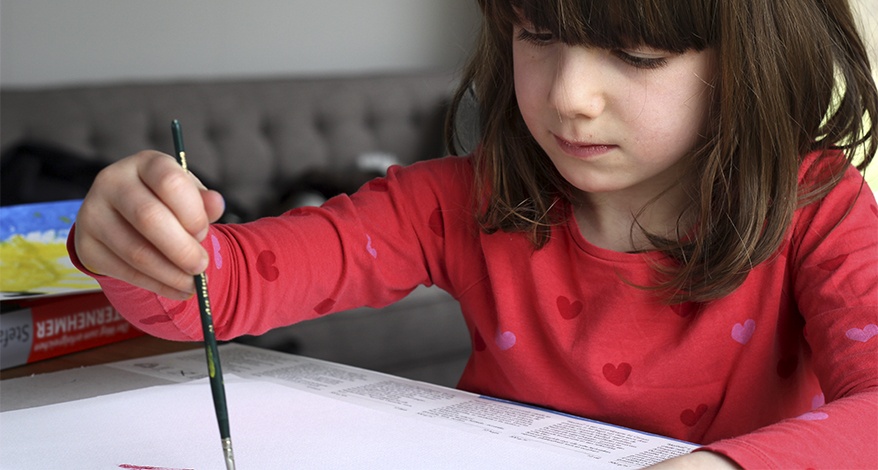 Ein Mädchen im Grundschul-Alter mit rotem Pullover sitzt an einem Tisch und halt in der rechten Hand einen Pinsel, mit dem sie etwas auf eine Schreibtischunterlage malt.