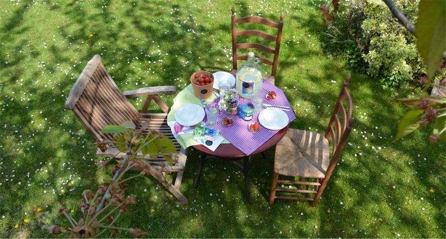 In einem Garten steht eine Sitzgruppe mit 3 Stühlen um einen bunt gedeckten Tisch. 