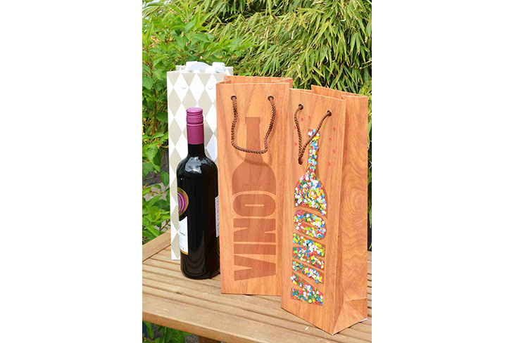 Auf einem Tisch im Garten stehen 3 Geschenktüten für Weinflaschen. Auf 2 Tüten ist der Schriftzug "Vino" zu lesen und es steht eine Flasche Rotwein vor einer Tüte.
