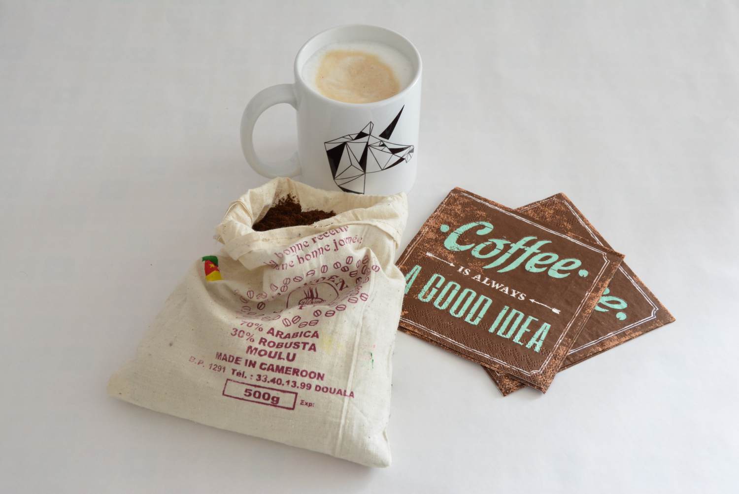 Auf einem Tisch steht eine Tasse Cappuccino, daneben liegen 2 braune Servietten mit der in grün geschriebenen Aufschrift "Coffee is always a good idea". Davor liegt ein Jutebeutel mit gemahlenem Kaffee.