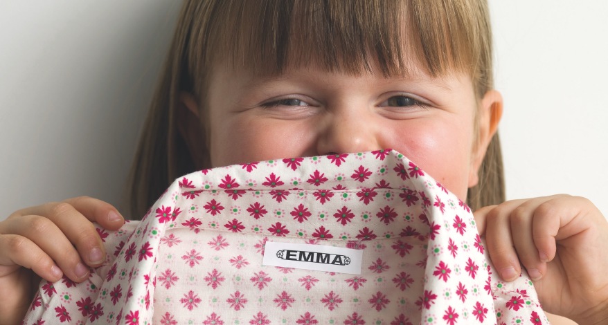 Ein Mädchen hält eine Bluse halb vor ihr Gesicht und lacht. Im Kragen der Bluse ist ein Schuld mit dem Namen Emma zu lesen.