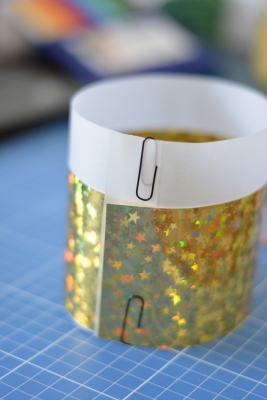 Ein Windlicht ist mit transparentem und goldenem Papier umwickelt und wird mit Büroklammern zusammengehalten.