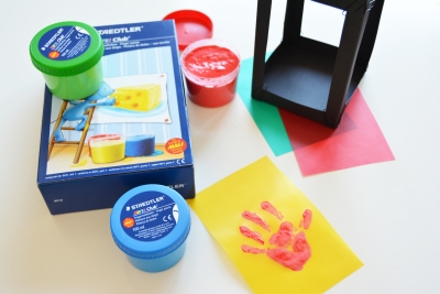 Auf einem Tisch steht eine Box der Marke Staedtler mit Finderfarben. 2 Töpfe der Farben stehen ausgepackt daneben. Es liegt ein gelbes Transpartentpapier mit einem roten Handabdruck auf dem Tisch und der Papierkorpus der Laterne steht daneben.