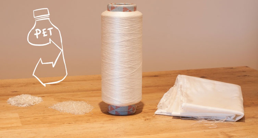 Auf einem Holztisch steht eine Spindel mit aufgewickeltem Garn, das aus PET hergestellt wurde. Daneben liegt ein zusammengefaltetes Stück Stoff und neben der Spindel liegen 2 kleine Haufen zerkleinertes Plastik. Auf das Bild ist eine Grafik einer PET-Flasche mit Recycling-Symbil gezeichnet.