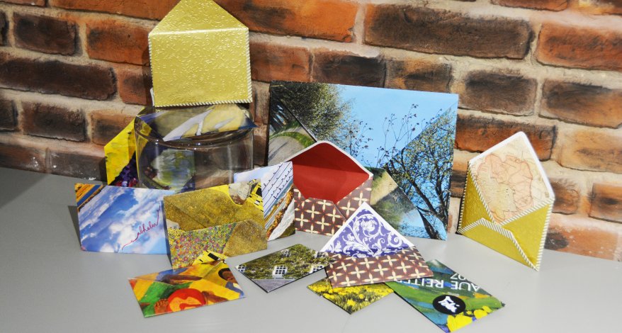 Auf einem Tisch vor einer geklinkteren Wand stehen und liegen viele bunte selbstgebastelte Briefumschläge.