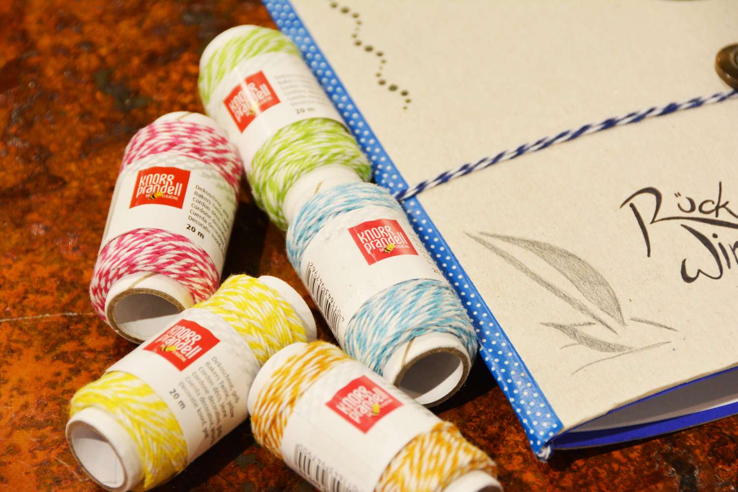 Ausschnitt eines selbstgebastelten Notizbuchs aus Karton. Daneben liegen diverse Rollen buntes Garn der Marke Heyda.