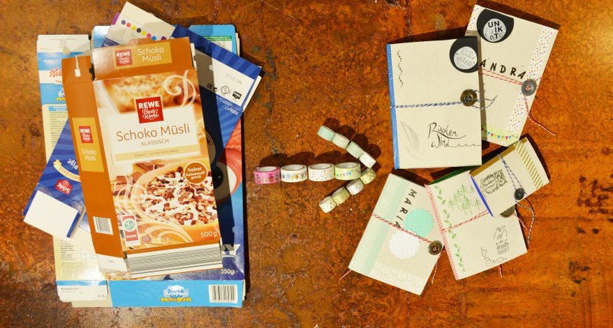 Auf einem Tisch liegen mehrere leere flach gefaltete Müslikartons. Daneben liegen ein paar Rollen Washitape und die aus den Müslikartons selbst gebastelten Notizbücher.