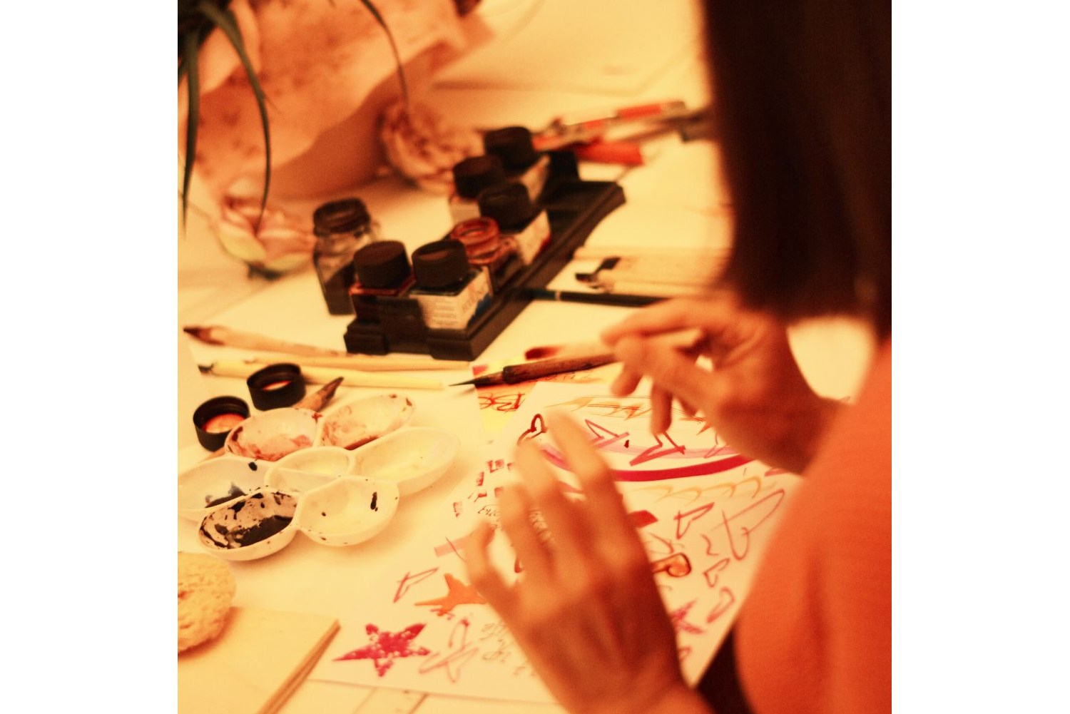 Vor einer Teilnehmerin des Kalligrafie-Kurses liegt ein Blatt mit roten Sternen und Herzen. Davor stehen mehrere bunte Tintenfässer und eine Schale mit Tinte.