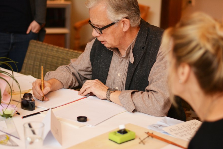 Kalligrafie-Experte Martin Heickmann sitzt am Tisch und schreibt mit einer Feder. Vor ihm steht ein Tintenfass und es liegt Papier auf dem Tisch.