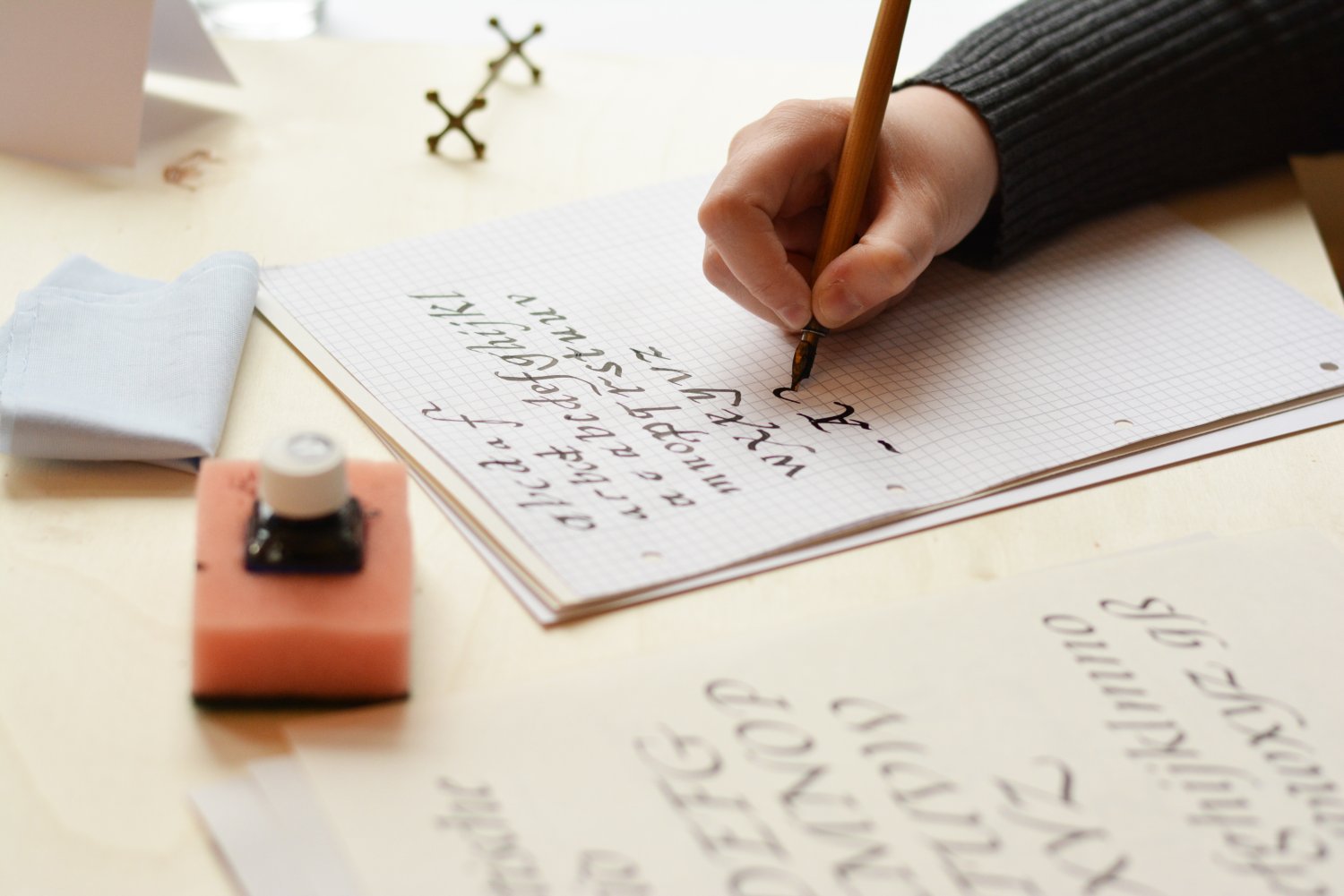 Auf einem Tisch liegt kariertes Papier, welches gerade mit Kalligrafieübungen beschrieben wird. Daneben liegen Vorlagen zur Orientierung. Vor dem Papier steht ein Fass mit Tinte.