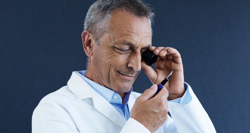 Mann im Laborkittel prüft einen Füllhalter mit einer Lupe und lächelt zufrieden.