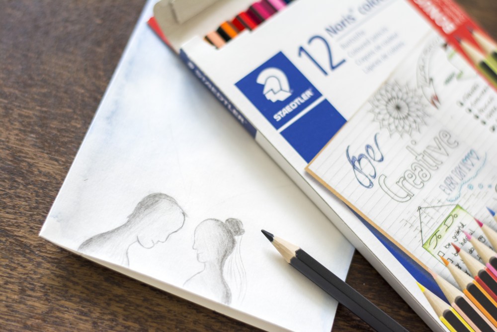Auf einem Holztisch liegt eine weiße Karte, am unteren Rand ist mit Bleistift ein Paar gemalt, das sich anschaut. Auf dem oberen Teil der Karte liegt eine  Packung Buntstifte der Marke Staedtler und ein blauer Buntstift.