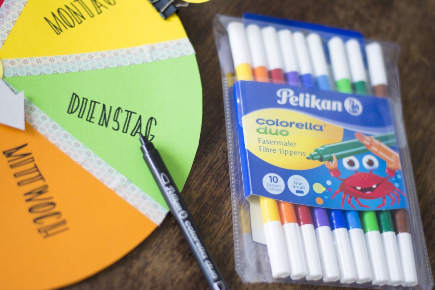 Auf einem Tisch liegt eine Packung Filzstifte der Marke Pelikan. Außerdem istdie Hälfte des selbstgebastelten Wochenplans mit den Tagen Montag, Dienstag und Mittwoch zu sehen.