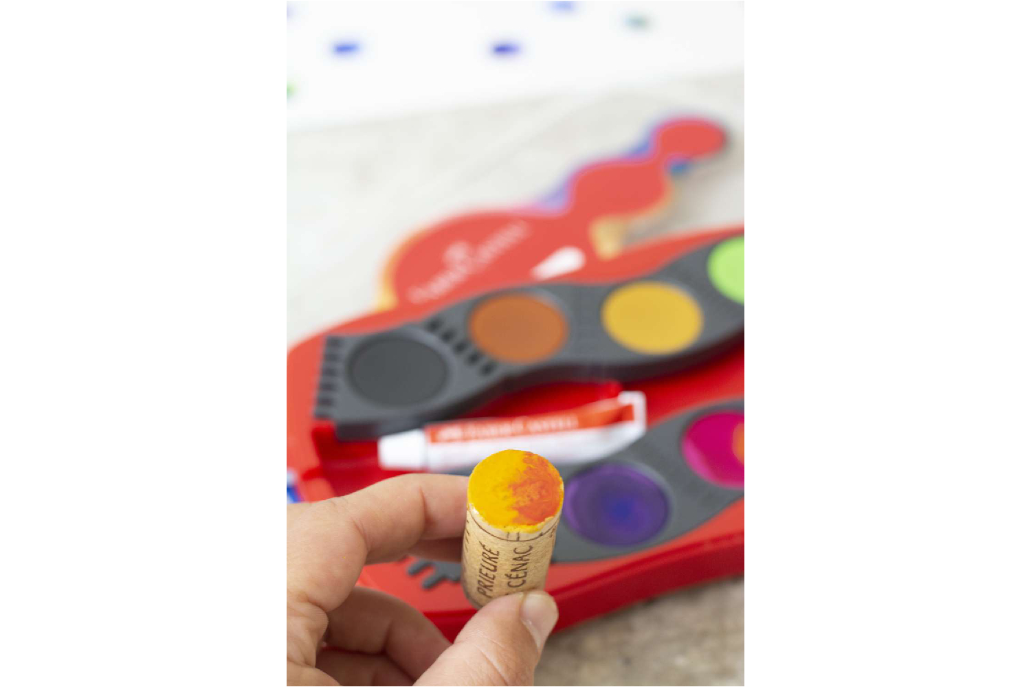 Eine Hand bestreicht Weinkorken mit blauer Farbe vor rotem Aquarellkasten