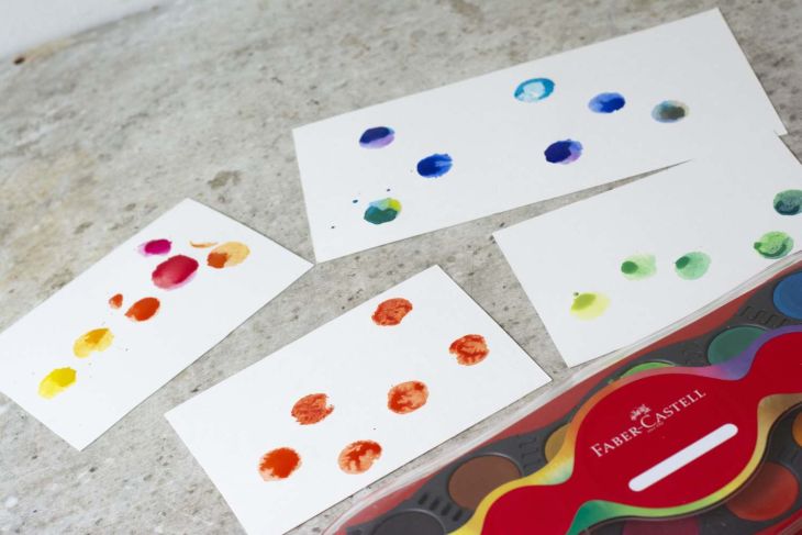 Mehrere Karten mit bunten Farbstempeln neben einem Farbkasten von Farber-Castell.