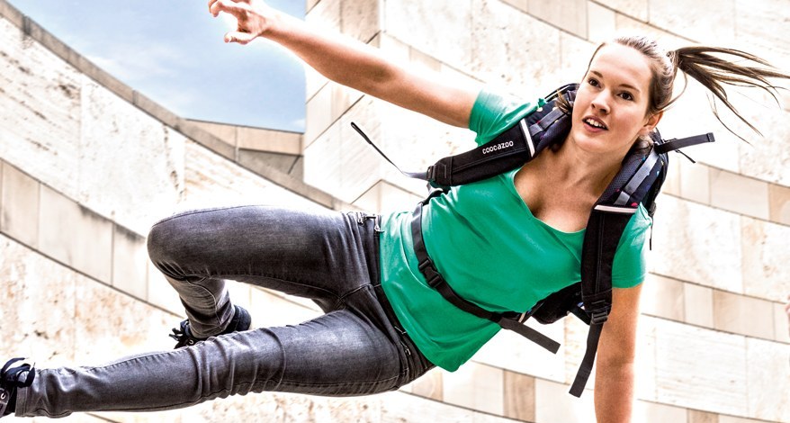 Ein jugendliches Mädchen springt über eine Mauer. Sie trägt eine schwarze Jeans, ein grünes T-Shirt und einen coocazoo Schulrucksack auf dem Rücken.