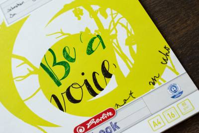 Auf einem gelben Schulheft mit Illustration der Marke herlitz ist der Spruch "be a voice" gelettert.