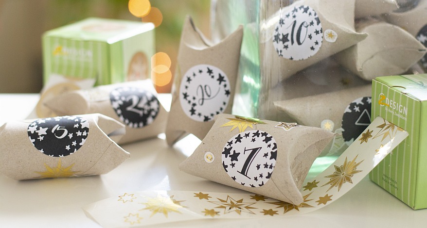 Klopapierrollen, die mit Nuimmern-Stickern beklebt sind und einen Adventskalender bilden. Daneben liegen die Verpackungen der Avery Zweckform Design Sticker.