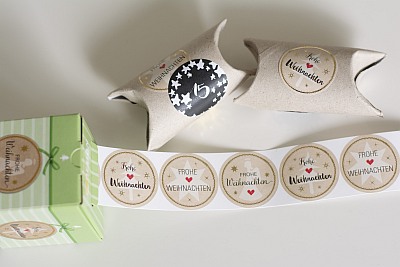 Weihnachtliche Sticker von der Rolle der Marke Avery Zweckform mit der Aufschrift "Frohe Weihnachten" liegen neben 2 beklebten Klopapierrollen.