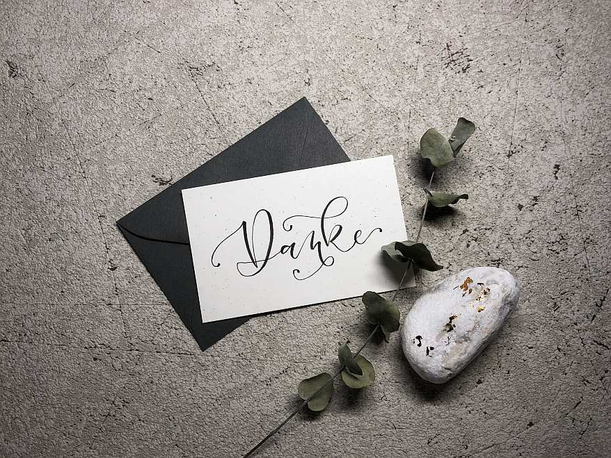 Auf einem Marmortisch liegt ein schwarzer Umschlag. Darauf liegt eine weiße Karte nmit einem schwarz gelettertem "Danke". Außerdem liegt als Deko ein Stein und ein Eukalyptusblatt daneben.