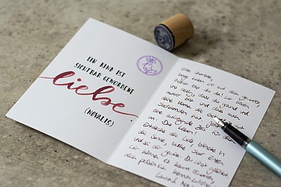 Eine aufgeklappte Karte, die auf der linken Innenseite mit einem Stempel und einem geletterten Sruch verziert wurde. Auf der rechten Seite steht Text und es liegt ein Füller auf der Karte.