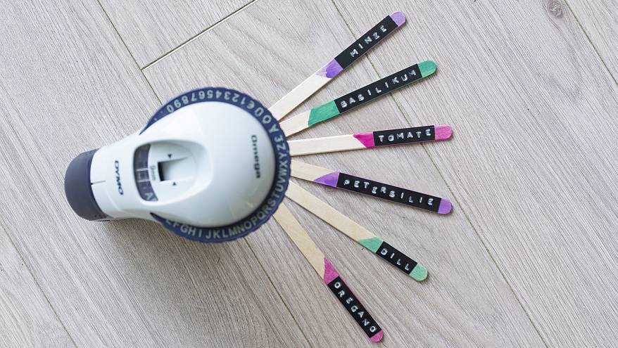 Auf einem weißen Parkettboden steht das Omega Prägegerät von Dymo. Darum verteilt liegen im Halbkreis verschiedene Eisstiele, die bemalt und mit Prägeband beschriftet wurden. Darauf sind verschiedene Namen von Kräutern zu lesen.