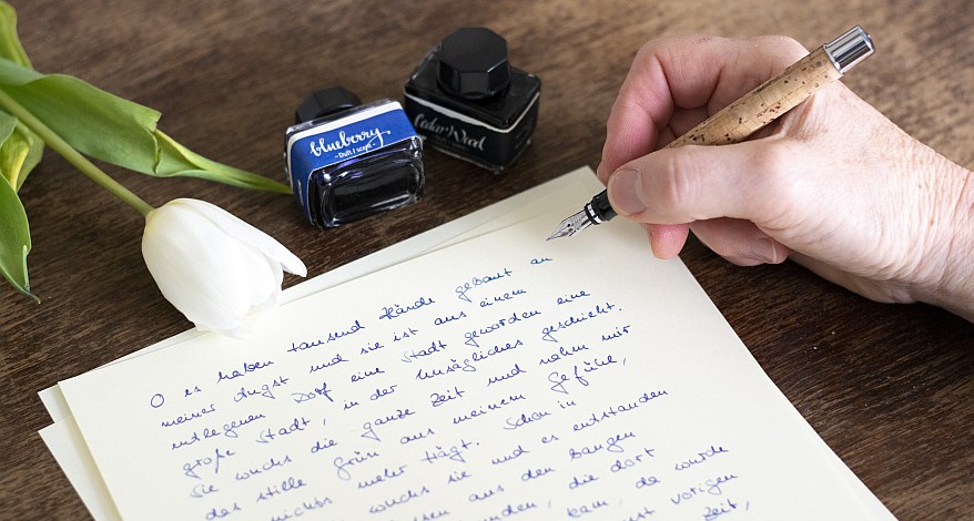 Hand schreibt mit Füller auf Briefpapier, daneben Tintenfass und eine weiße Tulpe.
