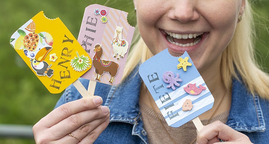Eine blonde Frau hält drei Einladungskarten in Form von Eis am Stiel in den Händen.