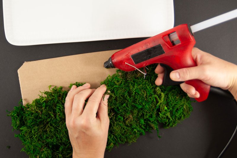 Ein Stück mit Moos wird mit Hilfe einer Heißklebefolie auf ein Stück Karton geklebt. Im Hintergrund steht eine weiße Schale. der Untergrund ist schwarz. 