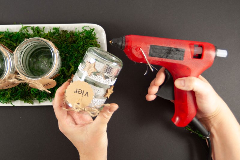 Auf ein verziertes Marmeladenglas wird mit Hilfe einer Heißklebepistole Kleber angebracht. Im Hintergrund ist ein Teil des DIY-Adventskalenders zu sehen. Auf Deko-Moos wurden bereits zwei fertige Gläser befestigt. Der Untergrund ist schwarz.